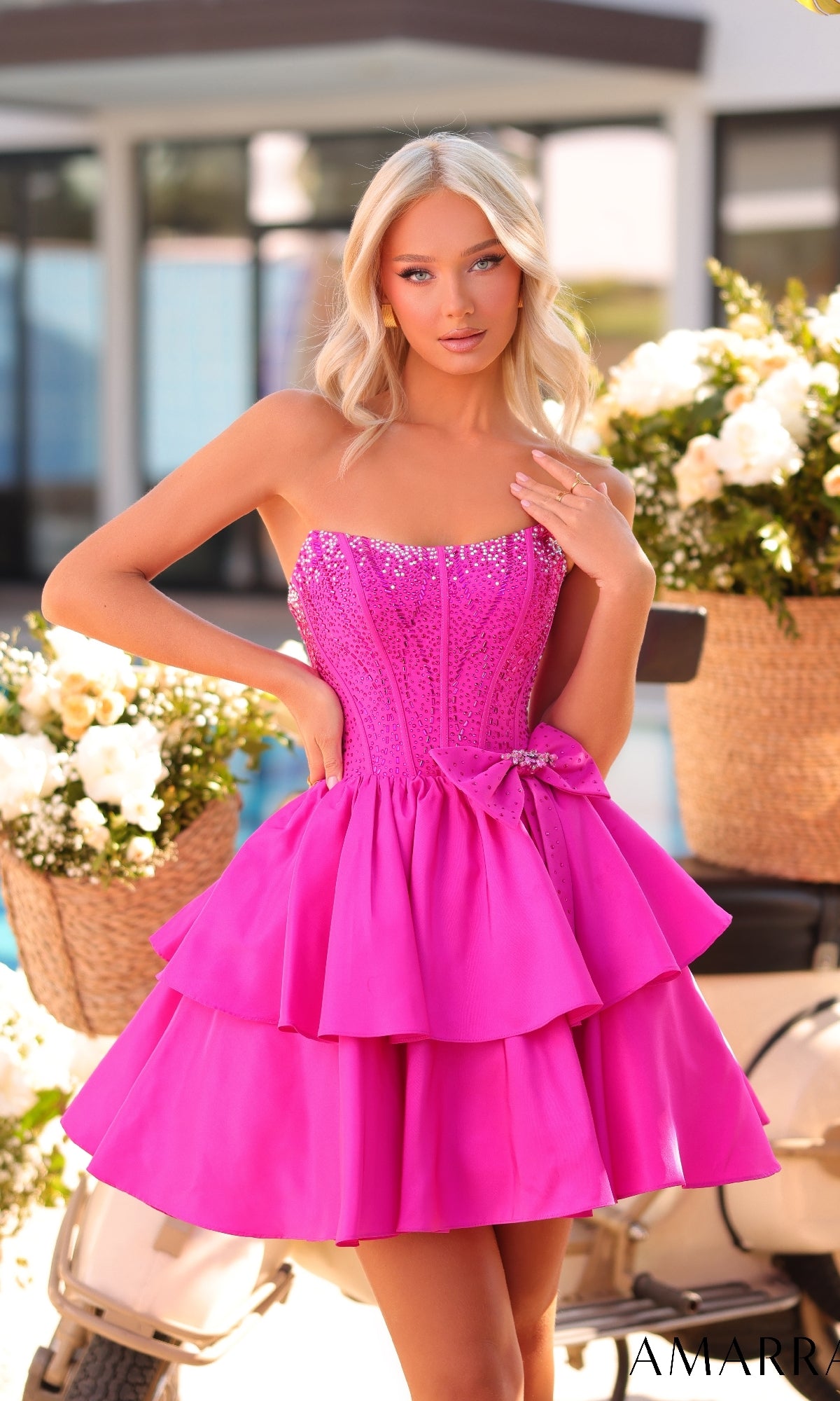A blonde woman stands outdoors by flowers and a scooter, wearing the Strapless Short Ruffled Hoco Dress: Amarra 88912 with a bright pink, tiered skirt.